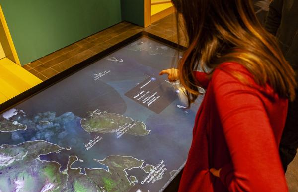 Two individuals stand over a touchscreen monitor tabletop. The woman is using her finger to tap the map on the screen.