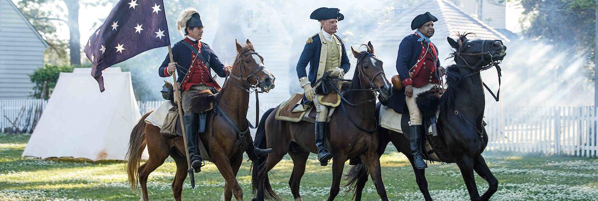 a reenactment of a Revolutionary War battle scene featuring 3 uniformed soldiers riding horses and carring a blue flag with white stars