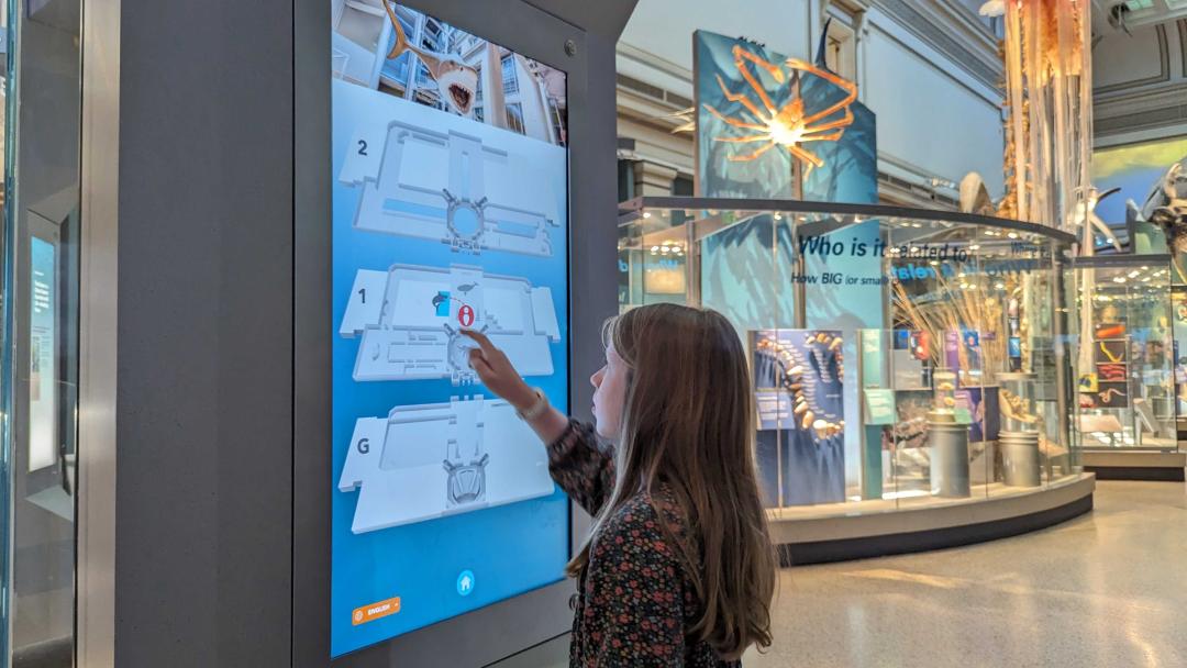 a young girl is touching a vertical mounted screen with floorplans displayed within a large exhibit hall